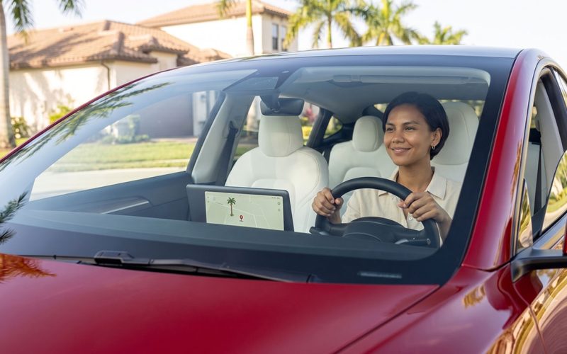 Smiling woman drives a red car with hands on the steering wheel and a tablet displaying a map on the dashboard in a sunny suburban street setting.